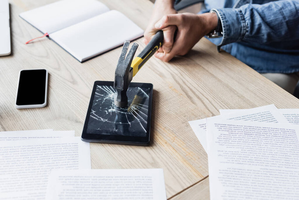 Vista cortada de empresário batendo tablet digital com martelo enquanto sentado no local de trabalho com documentos - Foto, Imagem
