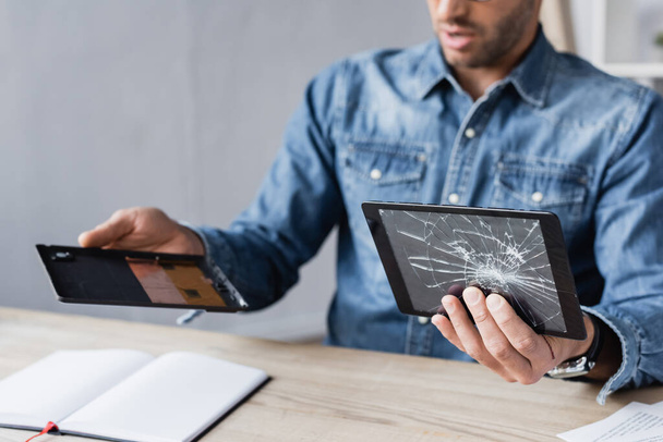 Gewassen beeld van zakenman demonteren van digitale tablet en het houden van gebroken touchscreen aan tafel op wazige achtergrond - Foto, afbeelding