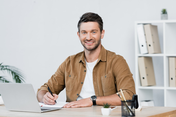 Uomo d'affari sorridente che guarda la fotocamera mentre scrive nel notebook sul posto di lavoro con laptop su sfondo sfocato - Foto, immagini