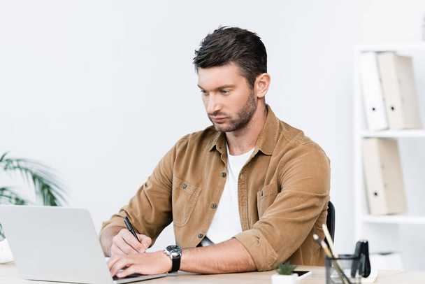 Focused businessman with pen looking at laptop while sitting at workplace on blurred background - Foto, Imagem