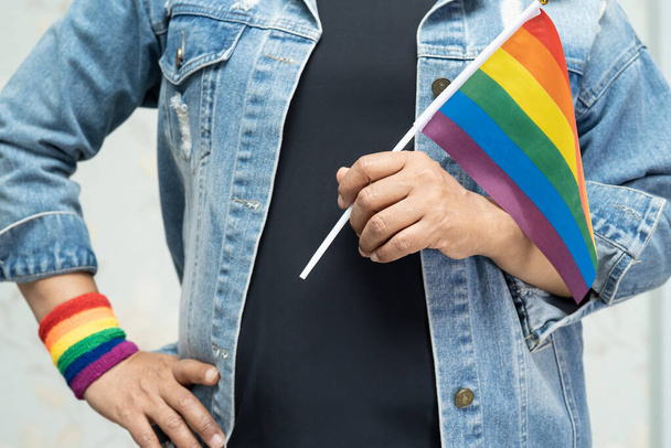 ゲイ、レズビアン、バイセクシャル、トランスジェンダー、人権の6月の社会でゲイ、レズビアン、バイセクシャル、トランスジェンダー、人権の毎年恒例のLGBTプライド月間のシンボルである、青いジーンジャケットやデニムシャツを着て虹色の旗を持っているアジアの女性. - 写真・画像