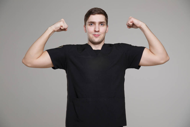 Young white man in casual working black clothes comically shows biceps isolated on a gray background. - Φωτογραφία, εικόνα