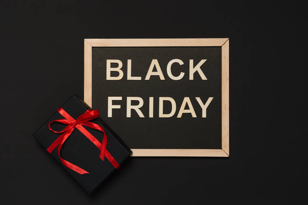 Black Friday sale concept. Black gift with red ribbon bow and blackboard with words from wooden letters on dark background. Flat lay style, top view - Photo, Image