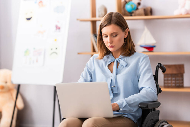 Laptopot használó női pszichológus kerekesszékben ülve, homályos irodával a háttérben - Fotó, kép