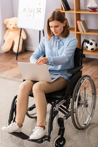 Toute la longueur de psychologue en utilisant un ordinateur portable tout en étant assis en fauteuil roulant dans le bureau - Photo, image