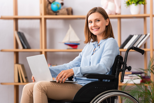 Happy female psychologist with laptop looking at camera while sitting in wheelchair on blurred background - Φωτογραφία, εικόνα