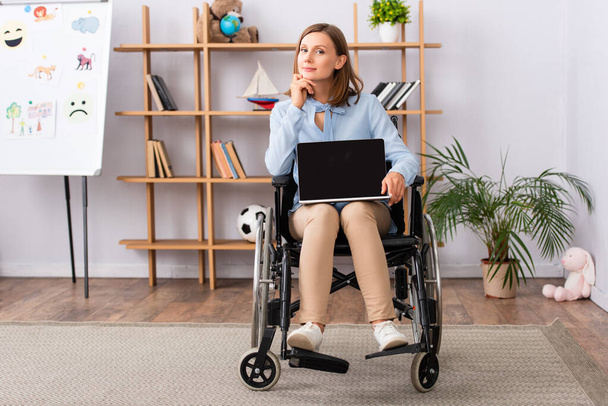 Full length of positive female psychologist showing laptop with blank screen while sitting in wheelchair in office  - Φωτογραφία, εικόνα