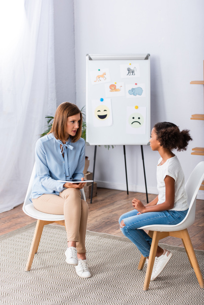 Full length of female psychologist with digital tablet talking to african american girl near flipchart in office - Photo, Image