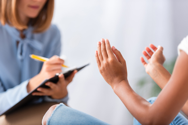 Vista recortada de niña afroamericana haciendo gestos cerca de psicólogo escribiendo en portapapeles durante consulta sobre fondo borroso - Foto, Imagen