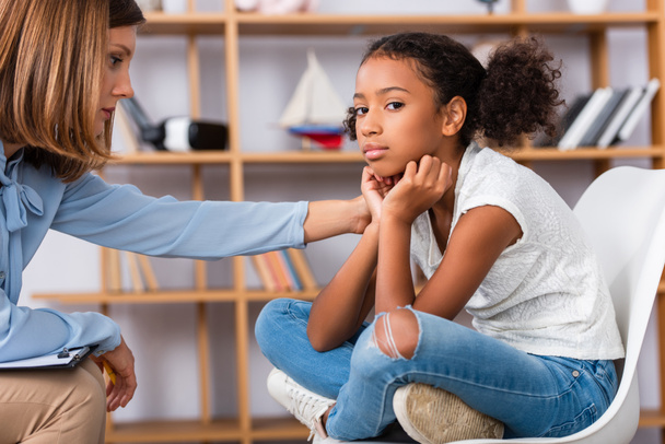 Psicólogo tocando hombro de niña afroamericana molesta mirando a la cámara durante la consulta sobre fondo borroso - Foto, imagen