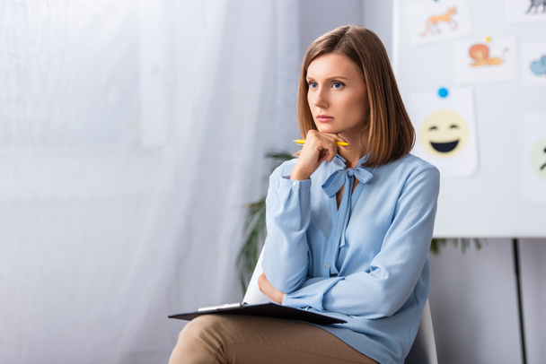 Psychologue féminine réfléchie avec stylo et presse-papiers regardant loin tout en étant assis sur une chaise avec un bureau flou sur fond - Photo, image