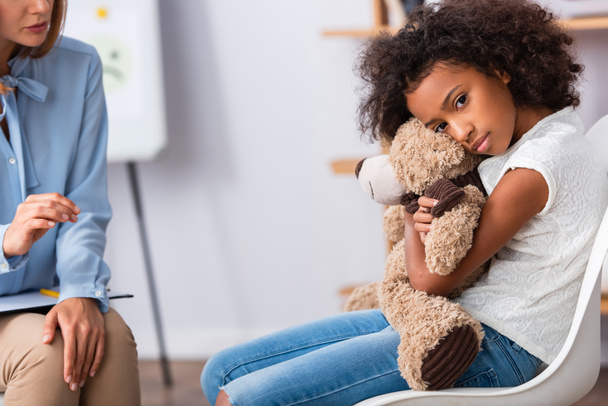 Depressed african american girl with autism looking at camera and hugging teddy bear near psychologist with blurred office on background - 写真・画像