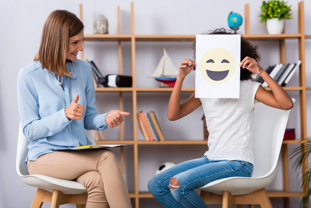 Smiling psychologist with thumbs up looking at african american girl covering face with happy expression on paper during consultation - Фото, зображення