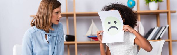 Menina afro-americana com autismo cobrindo rosto com expressão infeliz no papel perto psicólogo em fundo borrado, banner - Foto, Imagem