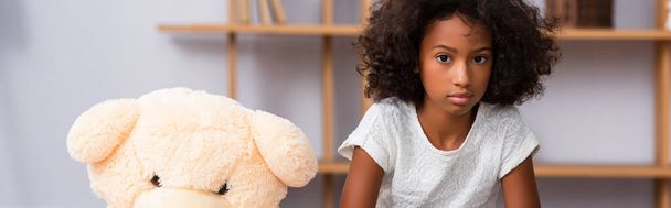 Upset african american girl with autism looking at camera near teddy bear with blurred office on background, banner - Fotoğraf, Görsel