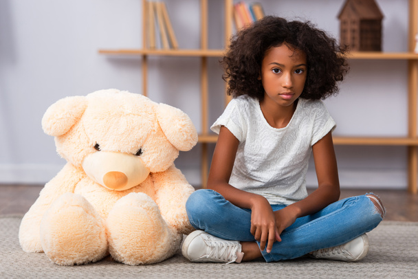 Depressed african american girl with crossed legs sitting on floor near teddy bear with blurred office on background - Fotó, kép