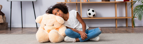 Depressed african american girl with autism looking away while leaning on teddy bear on floor near shelves in office, banner - Photo, Image