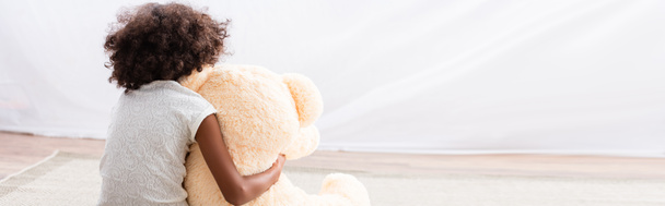 Yalnız Afrikalı Amerikalı kızın, yerde oyuncak ayıya sarılışının arkası. - Fotoğraf, Görsel
