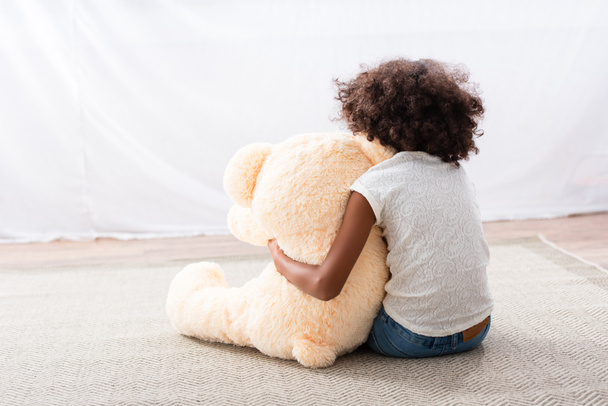 Rückansicht von einsamen afrikanisch-amerikanischen Mädchen mit Autismus umarmt Teddybär, während auf dem Boden in der Nähe Vorhang sitzen - Foto, Bild