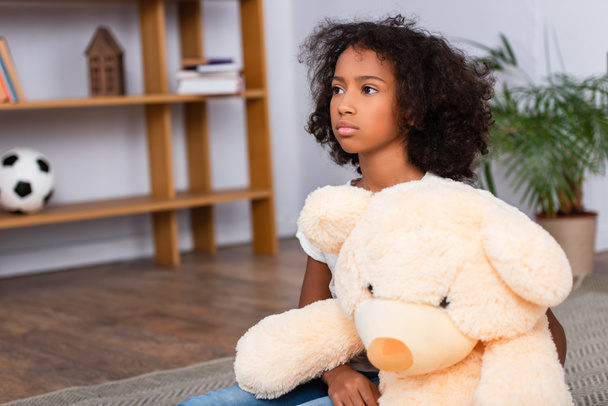 Verstoorde Afrikaans Amerikaans meisje weg te kijken terwijl knuffelen teddy beer op de vloer met wazig kantoor op de achtergrond - Foto, afbeelding
