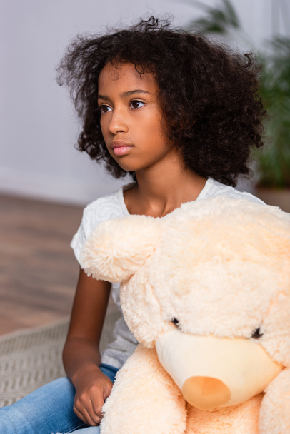 Пригнічена афро-американська дівчинка, яка дивиться на Тедді ведмедя на підлозі на розмитому фоні - Фото, зображення