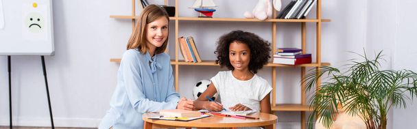 Happy psychologist and african american girl looking at camera near coffee table with colored pencils in office, banner - Фото, изображение
