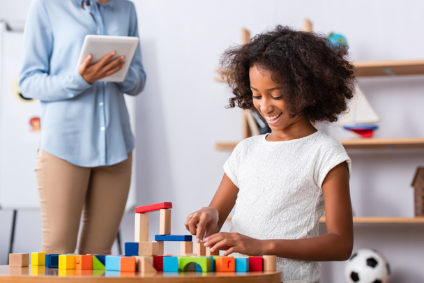 Happy african american girl playing with colorful wooden blocks at coffee table with blurred psychologist on background - Foto, immagini