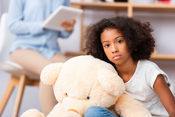 eenzaam afrikaans amerikaans meisje kijken naar camera in de buurt teddy beer met wazig psycholoog op de achtergrond - Foto, afbeelding