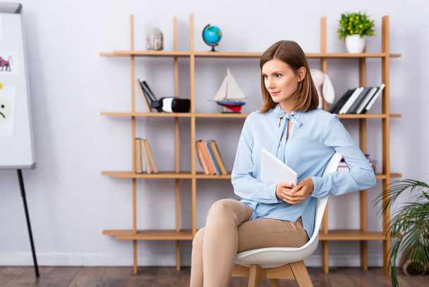 Жінка-психолог з цифровим планшетом дивиться вбік, сидячи на стільці в офісі
 - Фото, зображення