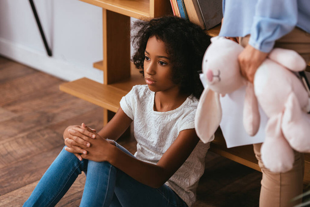 Depressed african american girl with autism looking away near psychologist standing near with soft toy on blurred foreground - Photo, Image