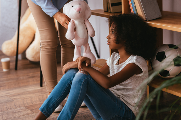 Upset african american girl with autism looking at soft toy in hands of psychologist standing near on blurred foreground - Фото, зображення