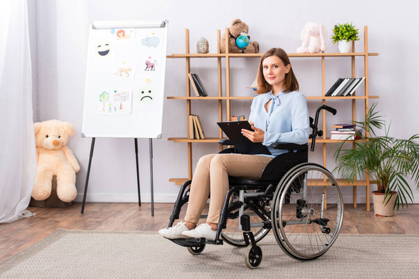 Volledige lengte van de positieve psycholoog met klembord kijken naar de camera tijdens het zitten in een rolstoel op kantoor - Foto, afbeelding