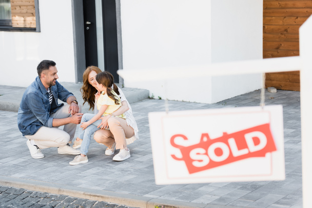 Счастливая семья приседает рядом с домом с размытым знаком с продаваемыми надписями на переднем плане - Фото, изображение