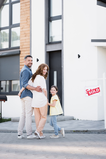 Полный рост улыбающейся семьи с дочкой, смотрящей на камеру, стоя возле дома, и табличка с проданной повязкой - Фото, изображение