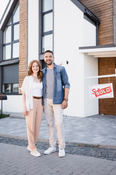 Full length of smiling couple hugging and looking at camera near sign with sold lettering and house - Foto, Imagen