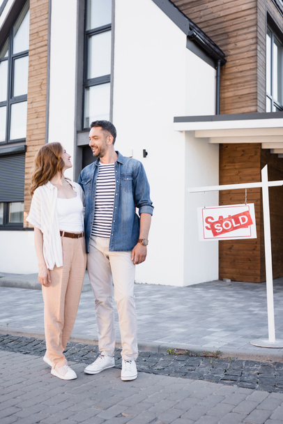 Full length of smiling couple looking at each other while hugging near house and sign with sold lettering - Foto, Imagem
