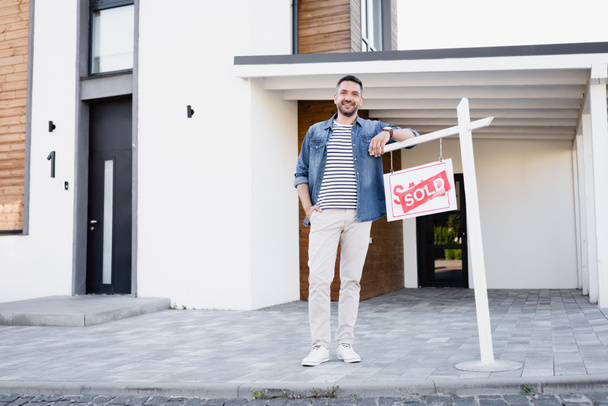 Full length of smiling man with hand in pocket leaning on sign with sold lettering near house - Photo, Image