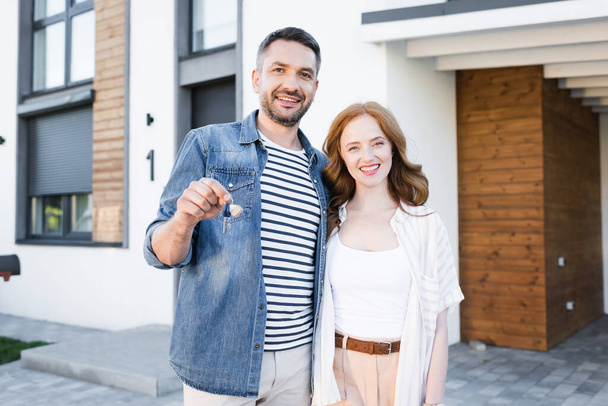 Happy man with key hugging woman while looking at camera with blurred house on background - Photo, Image