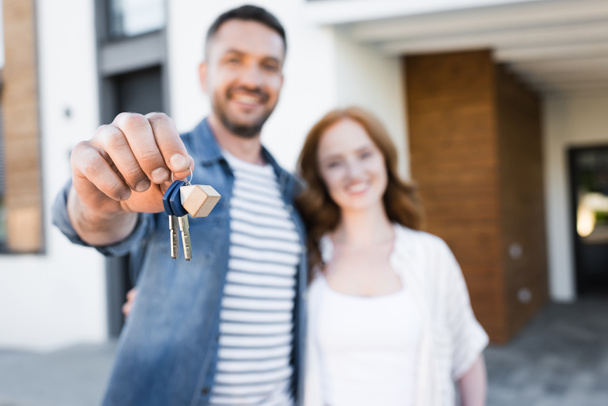 Κλειδιά και fob στο χέρι του ευτυχισμένος άνθρωπος αγκαλιάζει γυναίκα κοντά στο σπίτι σε θολή φόντο - Φωτογραφία, εικόνα