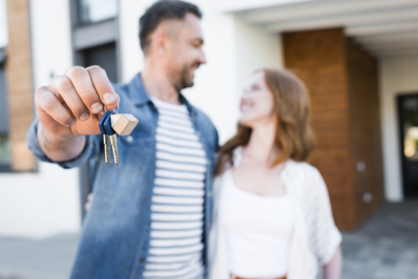 Ключи и брелок в руках счастливого мужа, смотрящего и обнимающего жену возле дома на размытом фоне - Фото, изображение