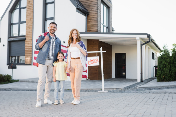 Longitud completa de hija feliz con mamá y papá sosteniendo la bandera americana mientras están de pie juntos y mirando la cámara cerca de la casa - Foto, Imagen