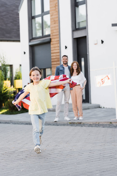 Täysi pituus onnellinen tyttö Amerikan lippu juoksee eteenpäin, kun katsot kameraa, jossa hämärtyneet vanhemmat halaavat taustalla - Valokuva, kuva
