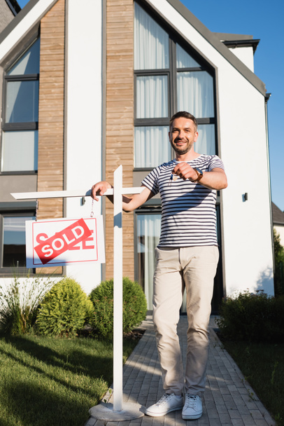 Täysi pituus hymyilevä mies osoittaa avain samalla seisoo lähellä merkki myydään kirjaimet moderni talo taustalla - Valokuva, kuva