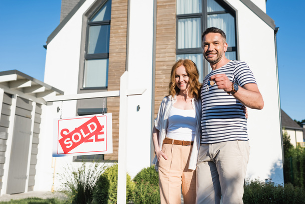 Smiling couple with keys hugging while standing near sign with sold lettering and modern house on background - Photo, Image
