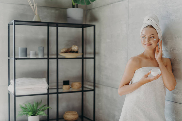 Photo of relaxed young woman applies face cream, has satisfied expression, wrapped in bath towel, poses near grey wall in bathroom, uses cosmetic product for rejuvenation. Beauty and health care - Фото, изображение