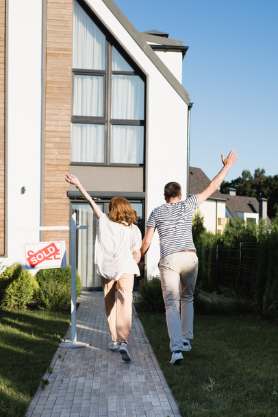 Vista trasera de pareja con las manos en el aire cerca de signo con letras vendidas y casa moderna - Foto, Imagen