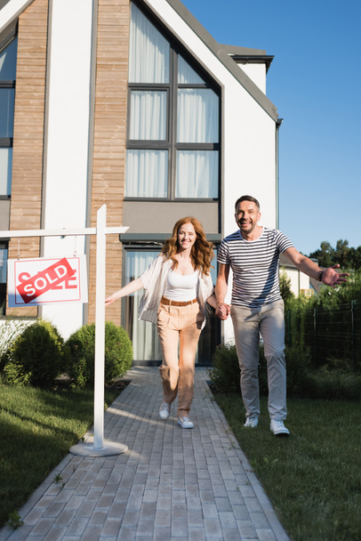 Cheerful couple holding hands while running forward near sign with sold lettering and modern house - Photo, Image