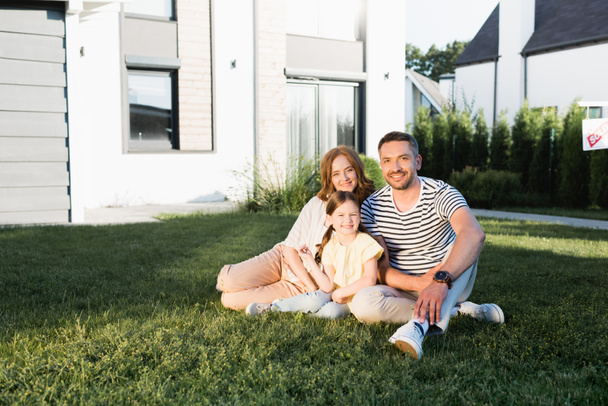 Glückliche Familie blickt in die Kamera, während sie auf dem Rasen neben modernem Haus sitzt - Foto, Bild
