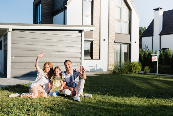Happy parents with waving hands and daughter sitting on lawn and looking at camera near house - Photo, Image