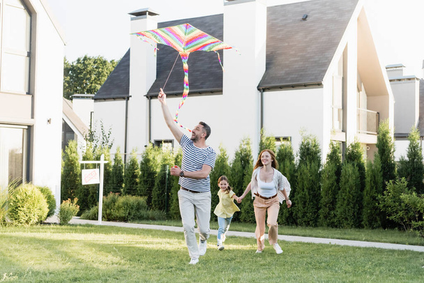 Full length of happy man flying kite near woman and girl on lawn near houses - Foto, Bild